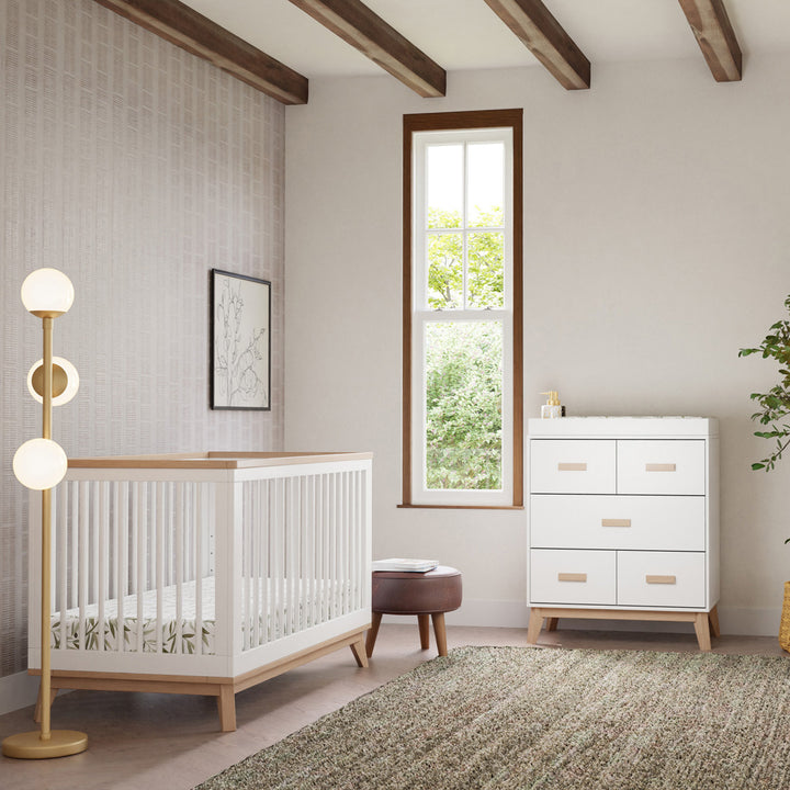 Babyletto's Scoot 3-in-1 Convertible Crib + Toddler Rail next to a lamp, stool, and dresser  in -- Color_Washed Natural/White