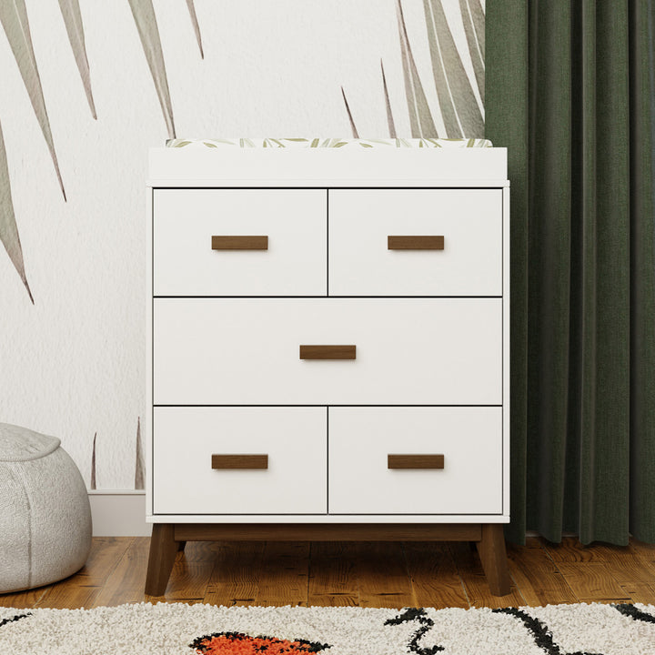 Front view of Babyletto's Scoot 3-Drawer Changer Dresser next to a green curtain in -- Color_White/Natural Walnut