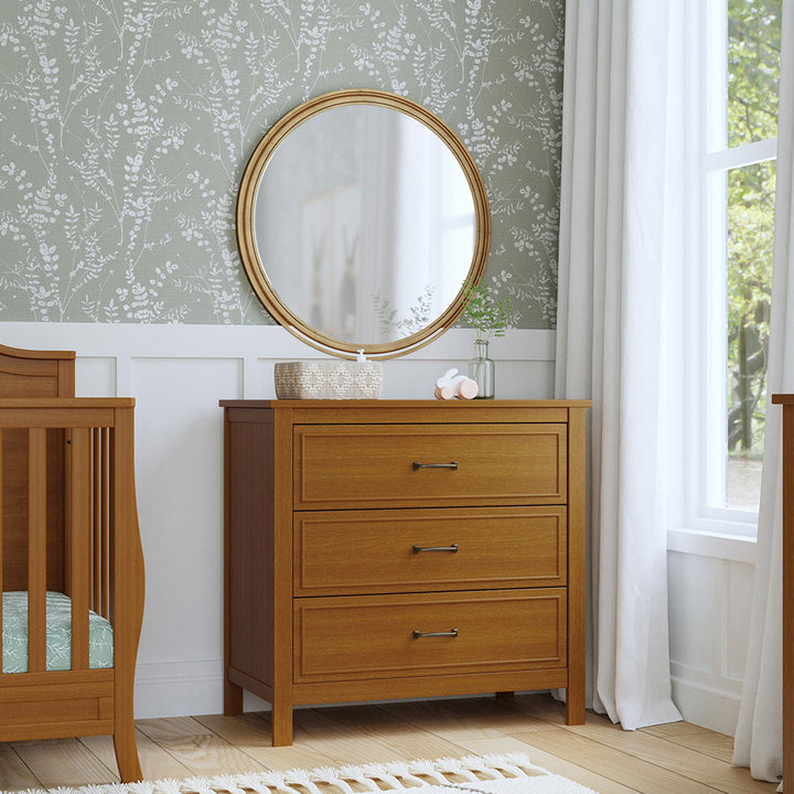 The DaVinci Charlie 3-Drawer Dresser under a mirror next to a crib  in -- Color_Chestnut