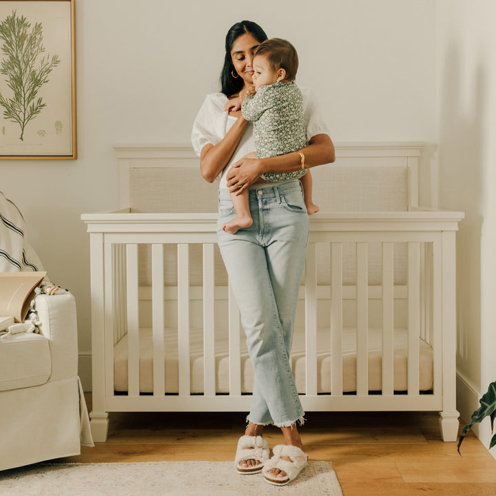 Mother holding a baby in front of the Darlington 4-in-1 Convertible Crib in Warm White