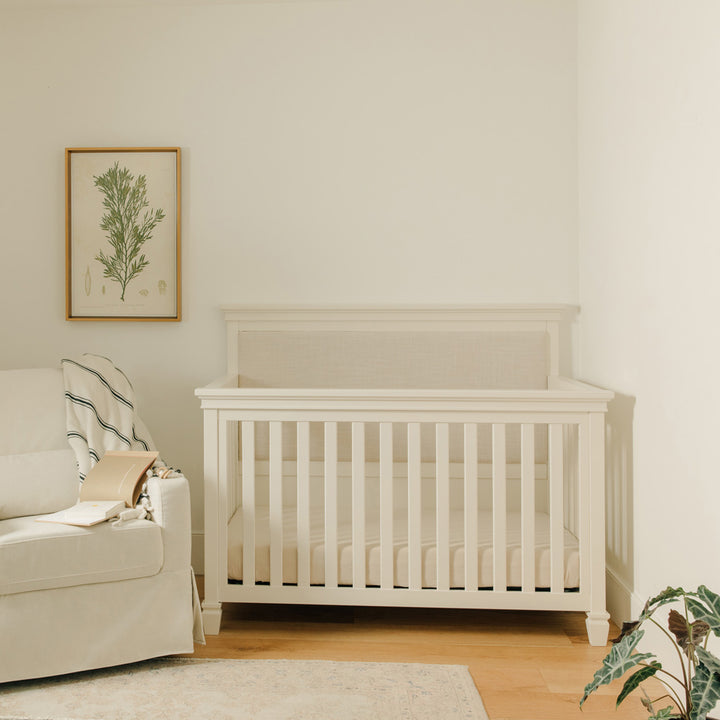 Darlington 4-in-1 Convertible Crib in Warm White next to a picture and a recliner