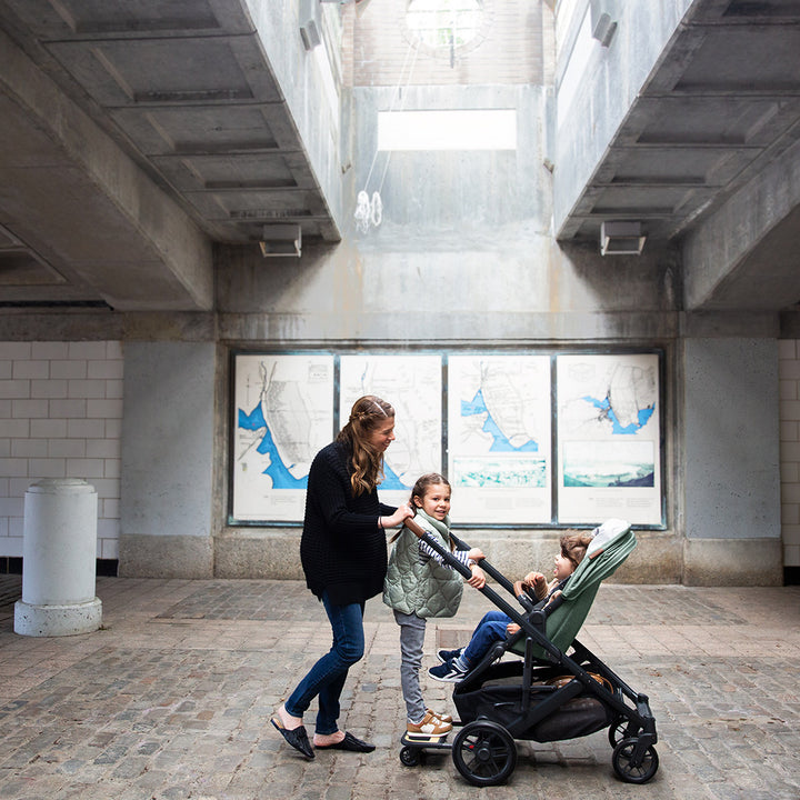 Side view of mom with a toddler pushing a baby in the UPPAbaby Cruz V2 Stroller in -- Color_Gwen