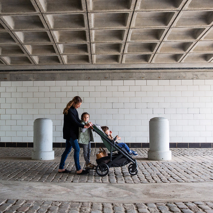 A mom with a toddler standing on the piggyback and a toddler sitting in the UPPAbaby CRUZ V2 Stroller in -- Color_Gwen