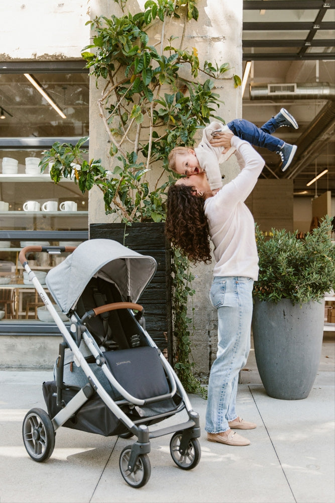 Mom holding up a toddler next to the UPPAbaby CRUZ V2 Stroller in -- Color_Gregory